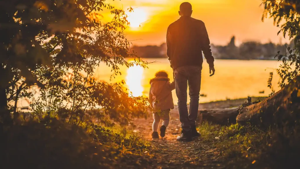 Silhouette of a man and child walking hand in hand near a lake at sunset.